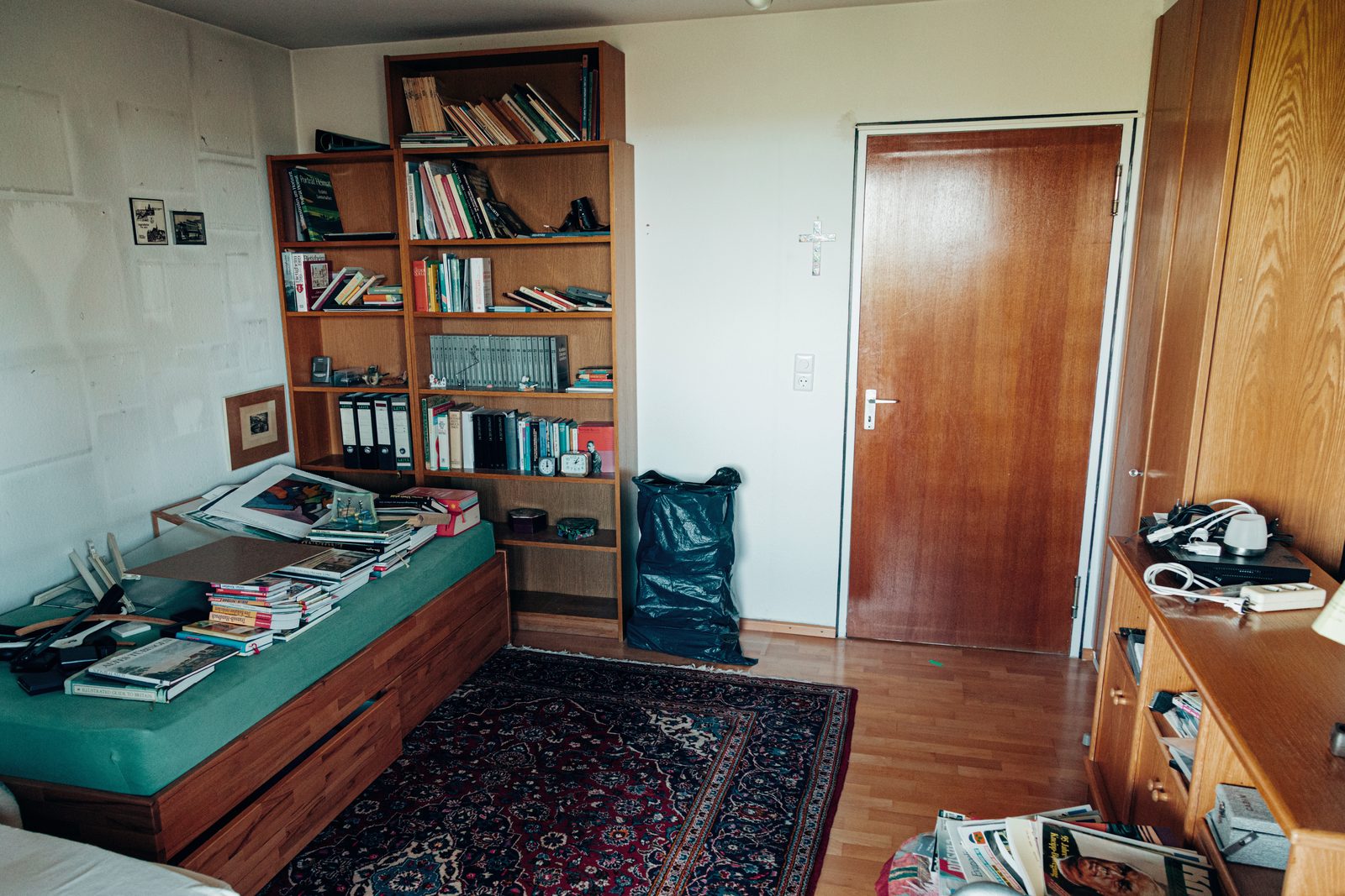 Arbeitszimmer vor der Räumung mit Bücherregal, Schlafsofa voller Bücher und Teppich.